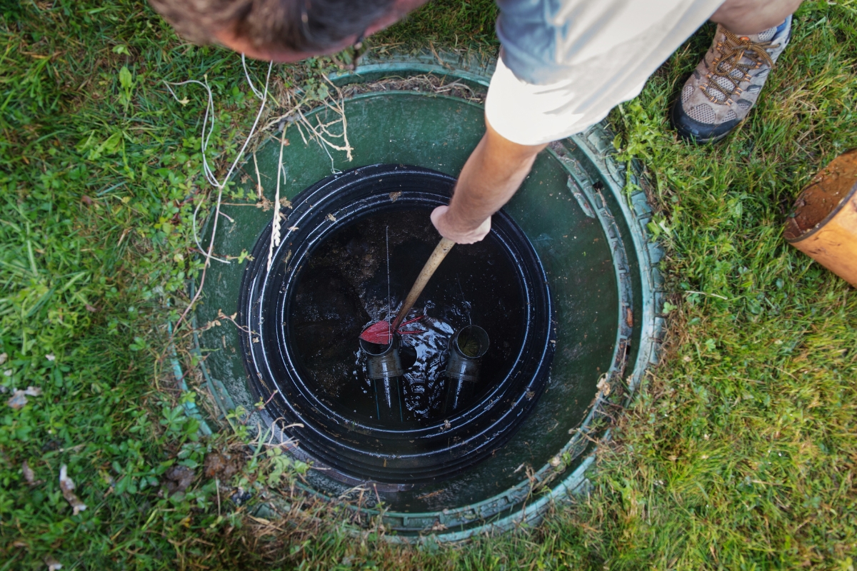 A person inspects and cleans a waterwell, with one hand holding a tool, standing on grass. The circular structure is partially filled with water, possibly indicating a septic tank backup.