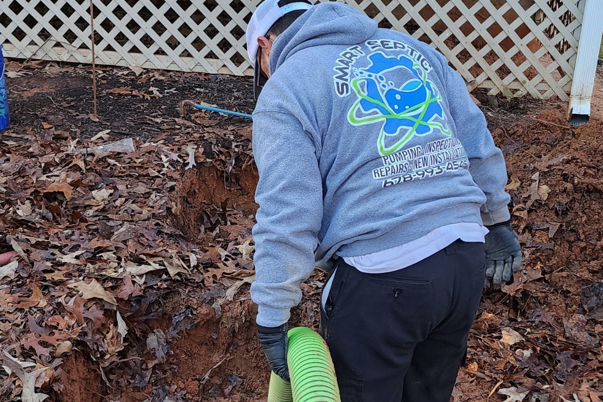 A worker in a gray hoodie with "Smart Septic" on the back handles a green hose, performing septic tank cleaning in a garden area with brown dirt and fallen leaves near white lattice fencing.