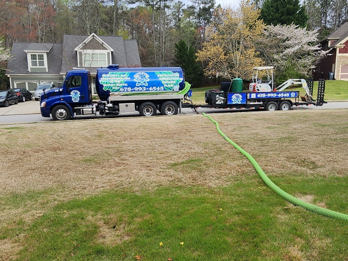 A septic service truck and trailer with hoses deployed in a residential neighborhood, parked on the side of the street near houses and trees, conducting a professional septic inspection and repair.