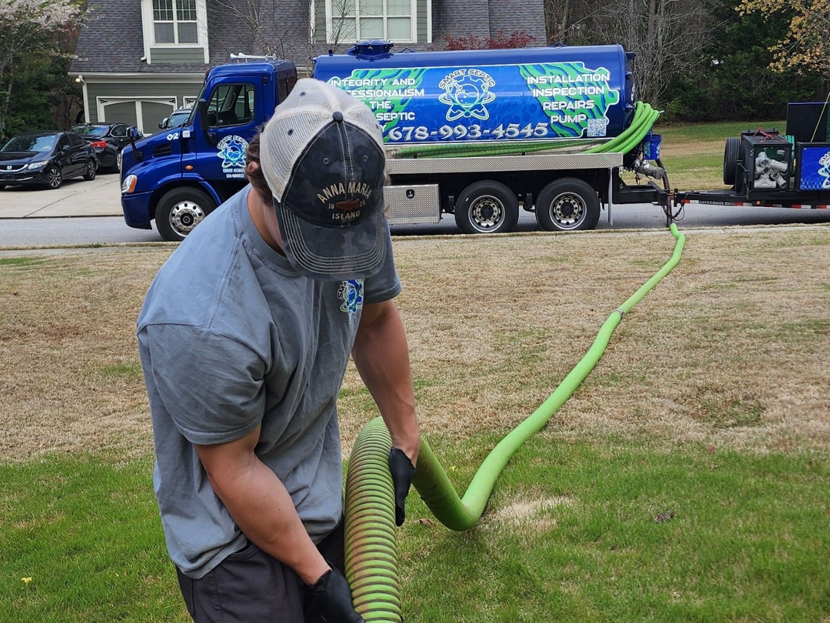 A Smart Septic Pros technician in a gray shirt and cap handles a green hose connected to a septic service truck parked on a residential street. The truck, advertising septic tank pump out services, displays a phone number for septic maintenance.