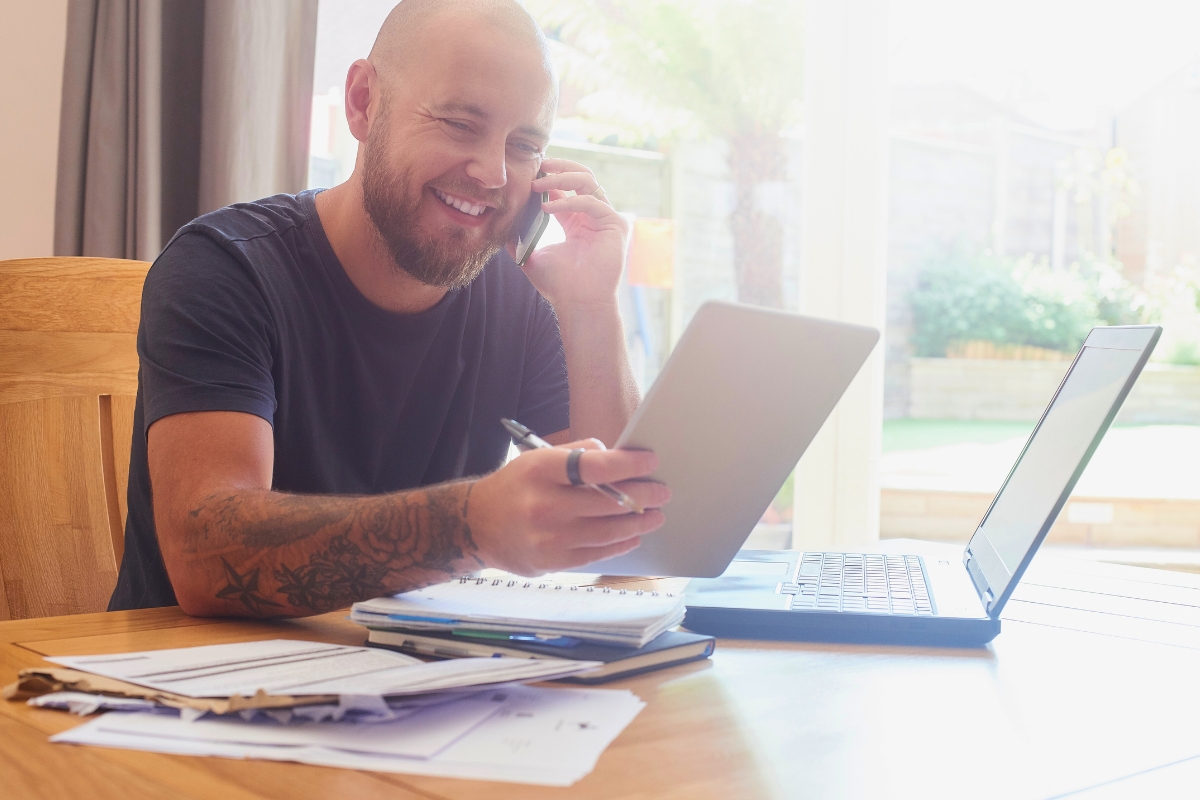 A man with tattoos sits at a table, using a laptop and holding a tablet while talking on the phone. Papers discussing septic tank pumping frequency and a notebook are spread on the table in a brightly lit room.