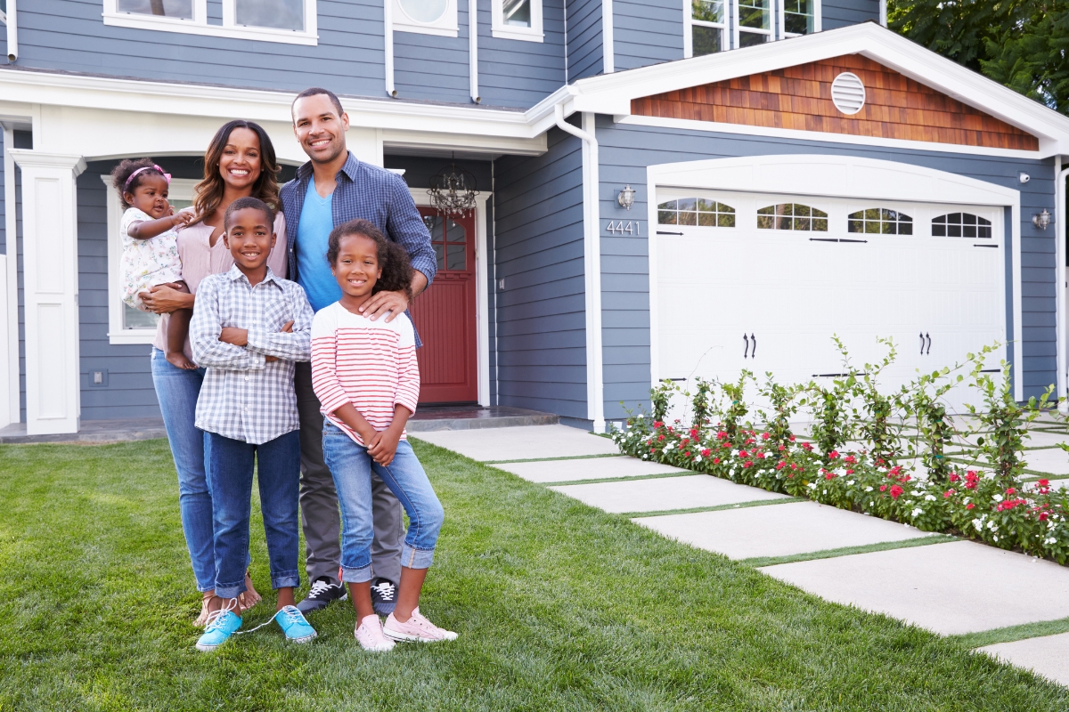 A family of four stands in front of their two-story blue house with white trim, smiling at the camera. The parents are holding a baby, and two children are standing in front of them, unaware that their home was thoughtfully built with ample septic tank sizes to accommodate their growing needs.