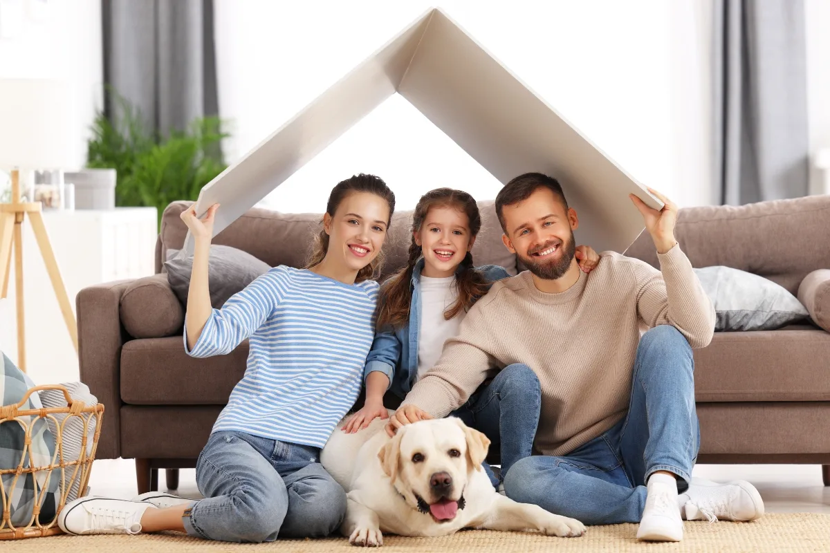 A smiling family of three sits in their living room with a Labrador. Two adults hold a makeshift roof structure over themselves and a child, symbolizing home-like warmth and protection, akin to how the right septic tank size ensures a safe and comfortable household environment.