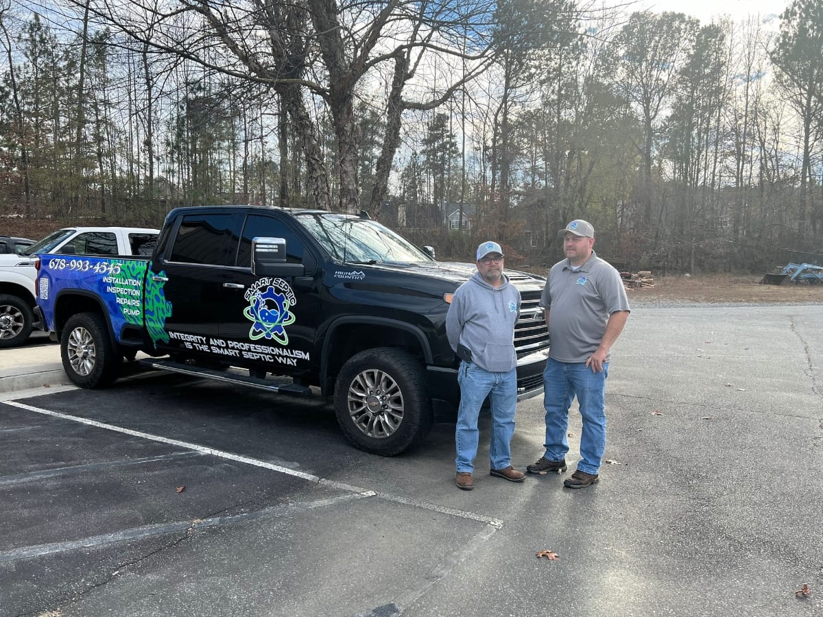 Two Smart Septic Pros standing beside a black truck with a company logo and contact information for septic cleaning costs, parked in a wooded area parking lot.