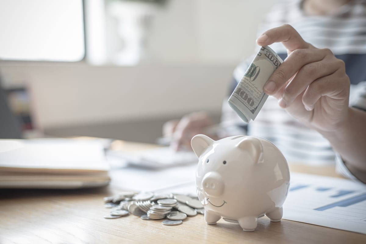 A person places a rolled-up banknote into a white piggy bank surrounded by coins on a table, much like investing in an aerobic septic system can bring long-term savings.