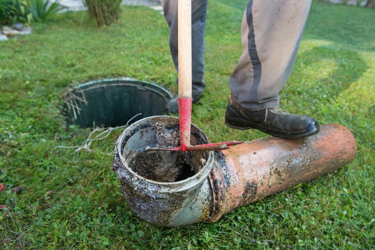 A person stands on the grass, using a tool to clean debris from a partially buried cylindrical drainage pipe connected to an aerobic septic system.