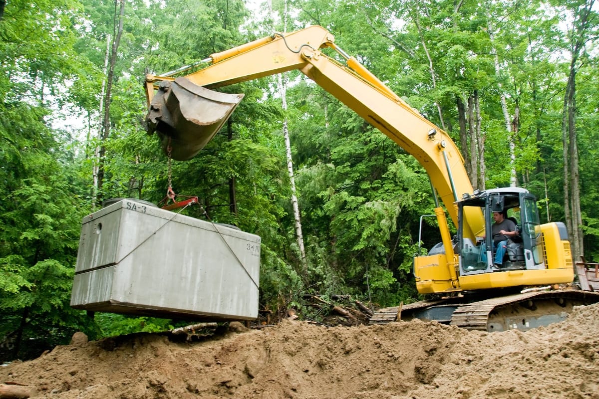 An excavator carefully lifts a large concrete block in a forested area, preparing the site for an aerobic septic system installation amidst the trees and dirt.