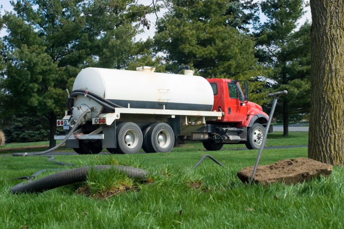 A red septic truck with a white tank, servicing an aerobic septic system, is parked on grass near trees. Hoses are extended onto the ground.