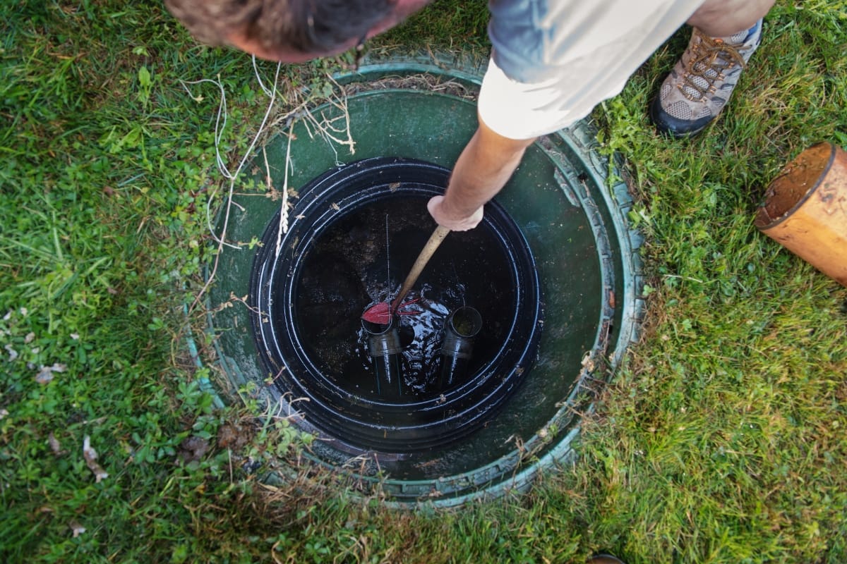 A person carefully inspects a round underground water meter with a long tool, surrounded by lush grass. The scene evokes the meticulous care required for maintaining an aerobic septic system, ensuring everything runs smoothly beneath the surface.