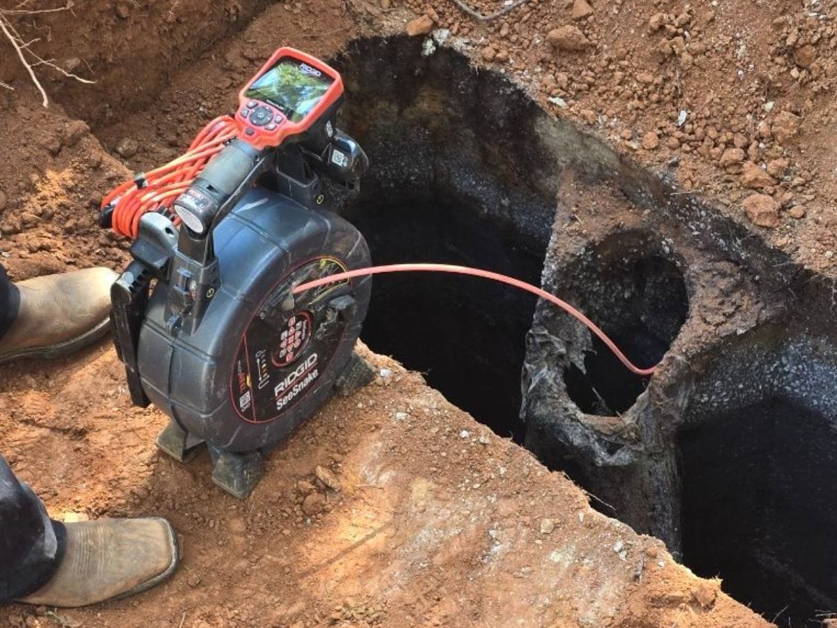 A device with a camera is used to inspect a pipe underground, ensuring compliance with septic inspection requirements. The camera cable extends into a dark hole surrounded by soil, while boots are visible near the setup.