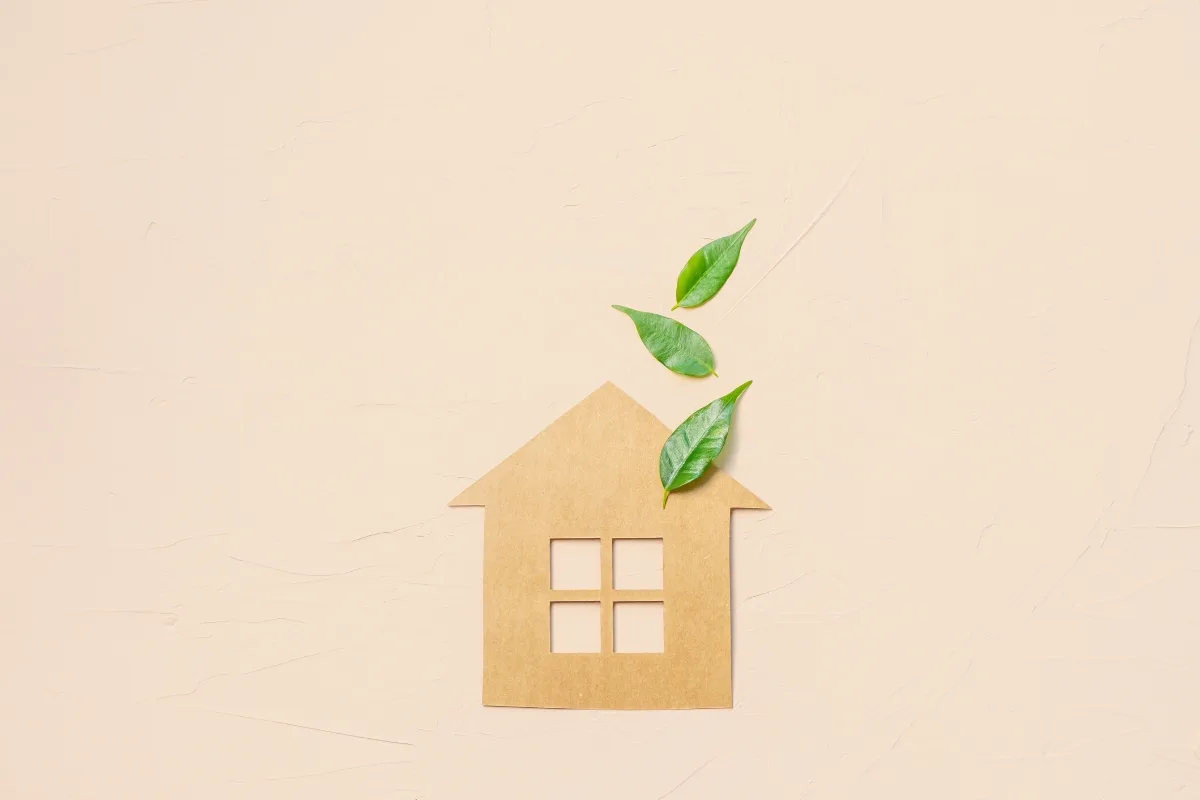 Cardboard house silhouette with window cutouts, paired with green leaves symbolizing nature’s harmony, on a beige background.