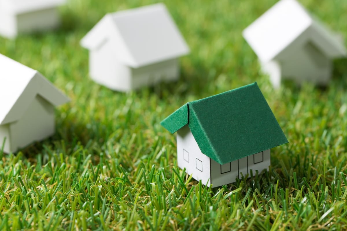 Small paper house models with green roofs are placed on a grassy surface, symbolizing sustainable living and echoing themes of green septic cleaning.