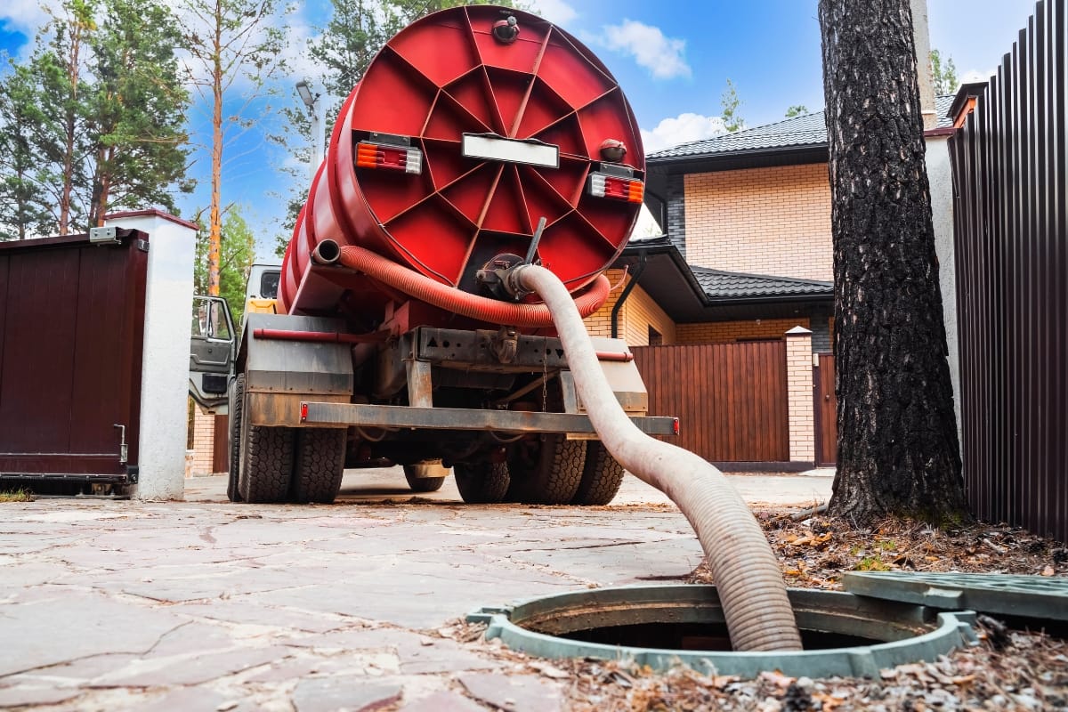 A red septic truck, emblazoned with "Green Septic Cleaning," is parked on a driveway. Its hose extends into an open septic tank, framed by trees and a house in the background.