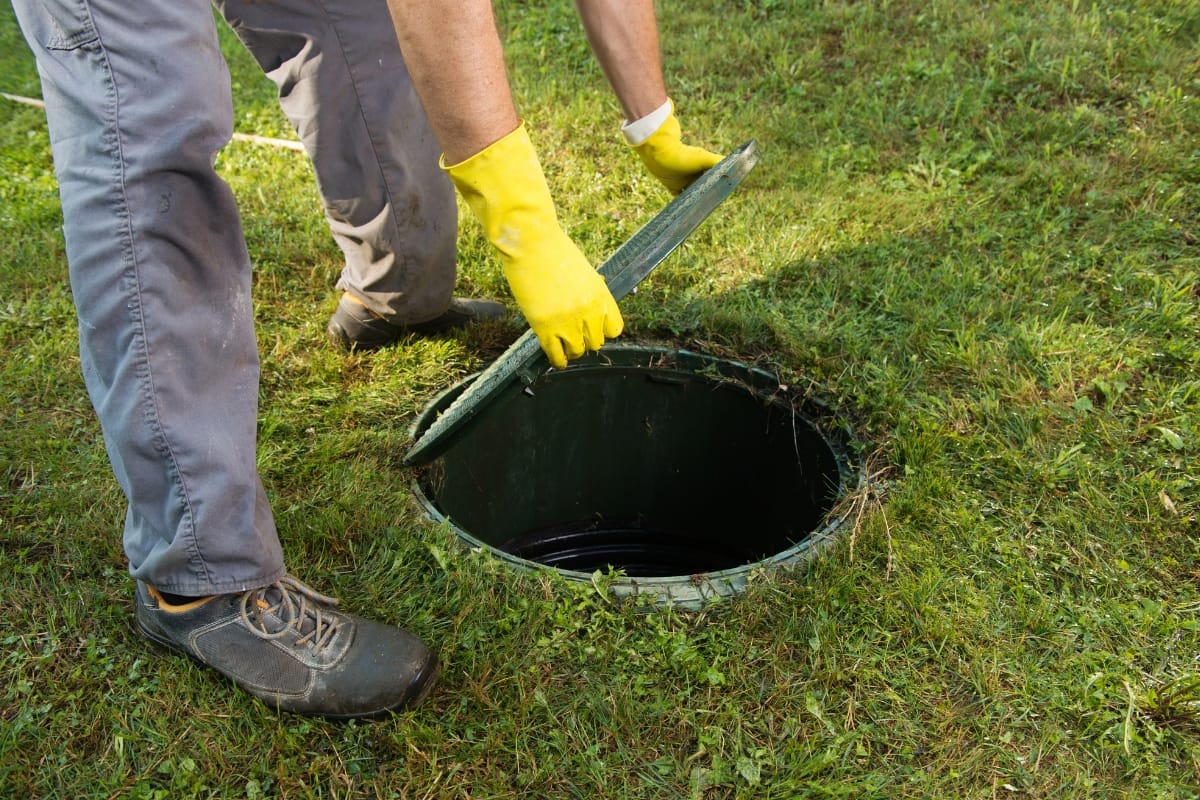 A person wearing yellow gloves carefully lifts the lid of a septic tank on a grassy lawn, showcasing their dedication to green septic cleaning.