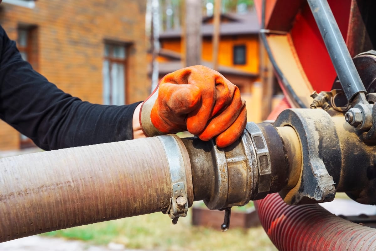 A person wearing orange gloves holds a large industrial hose attached to machinery, expertly demonstrating how often you should pump your septic tank, with buildings visible in the background.