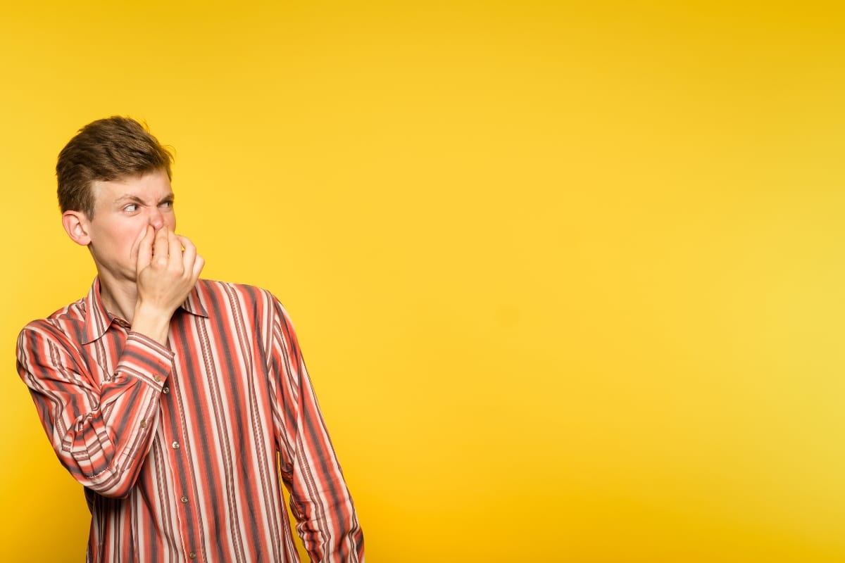 A man in a striped shirt pinches his nose with a disgusted expression, mimicking the unpleasant realization that can come when you learn how to know if your septic tank is full, all set against a bright yellow background.