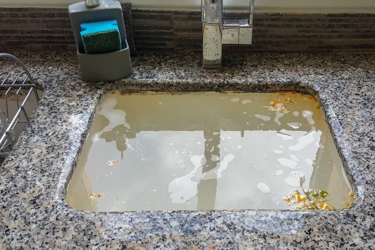 A kitchen sink filled with murky water, possibly hinting that it's time to check if your septic tank is full, contains food scraps and a sponge on the side with a faucet above.