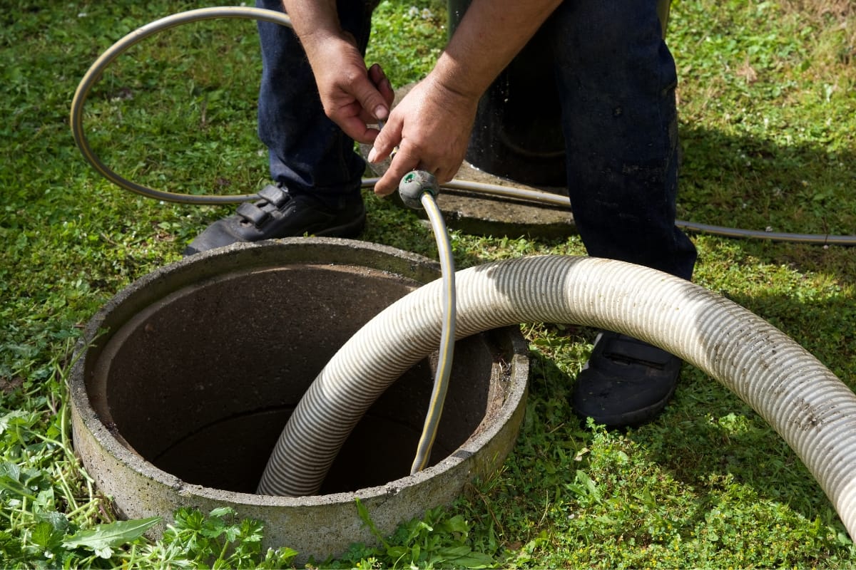 A person holding a hose near an open outdoor drain or septic tank, inspecting for signs that indicate how to know if your septic tank is full.