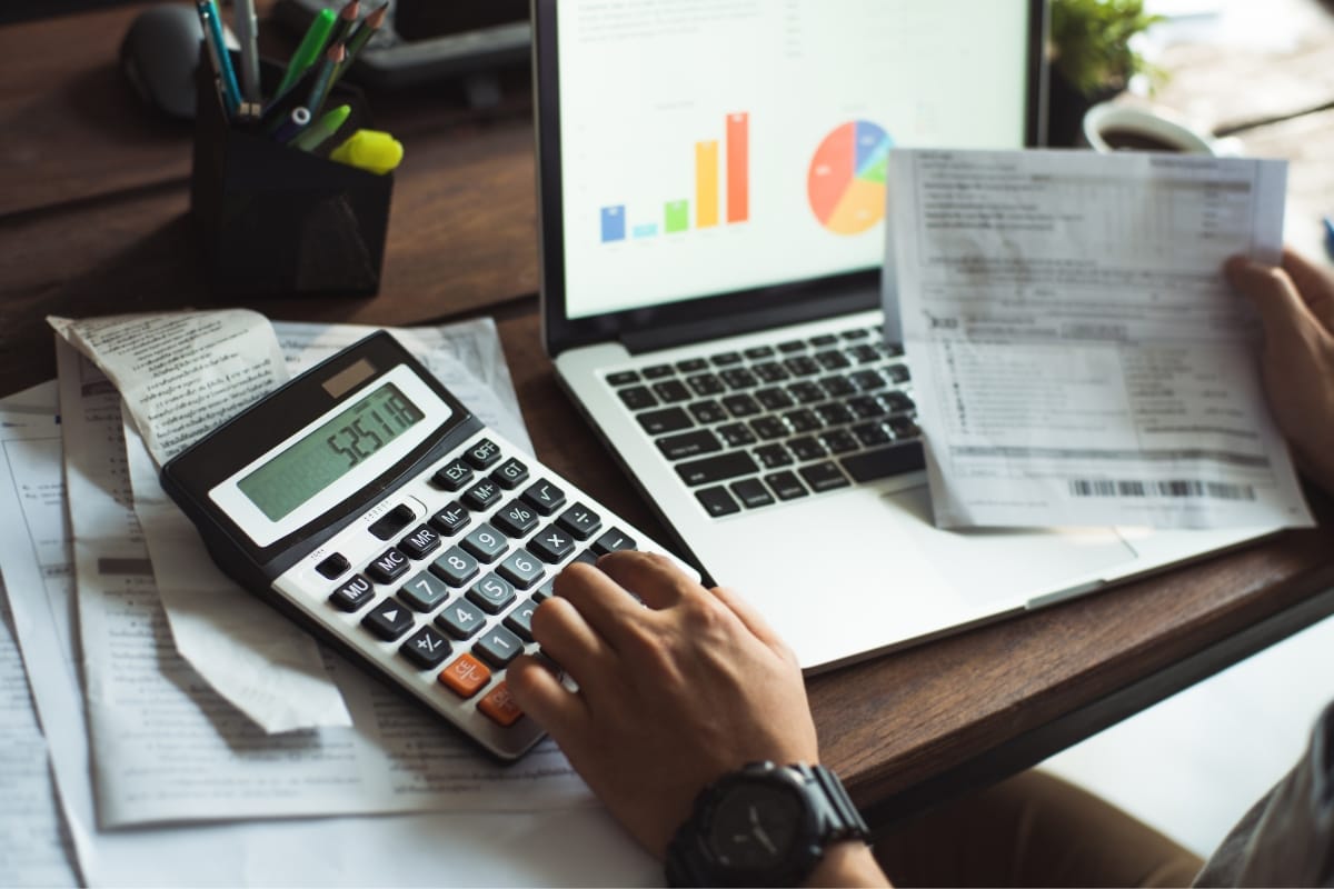 A person uses a calculator and reviews financial documents in front of a laptop displaying charts, comparing the costs and benefits of mound septic systems vs. conventional options.