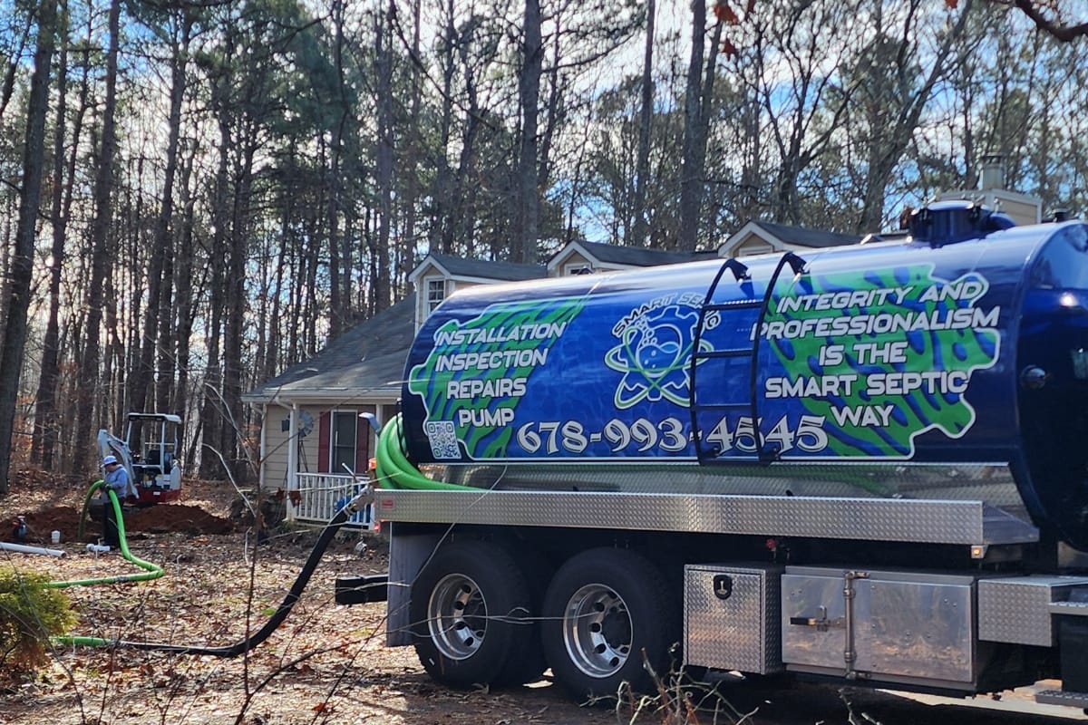 A septic service truck, its company details prominently displayed, is parked in front of a house with hoses extended. Surrounded by trees, the crew discusses mound septic system vs conventional options with the homeowner.