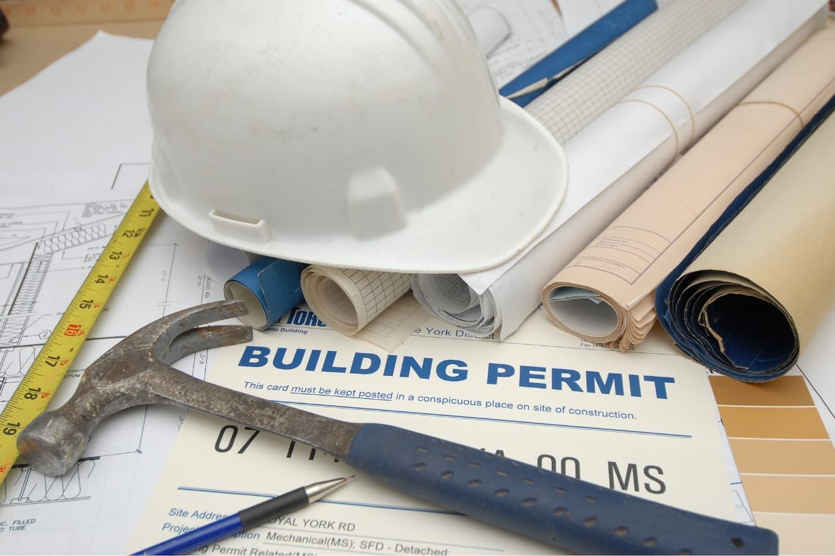 A white hard hat, rolled blueprints, a hammer, and a building permit are arranged on a table alongside a tape measure, illustrating the meticulous planning involved in construction projects like choosing between mound septic systems vs conventional ones.