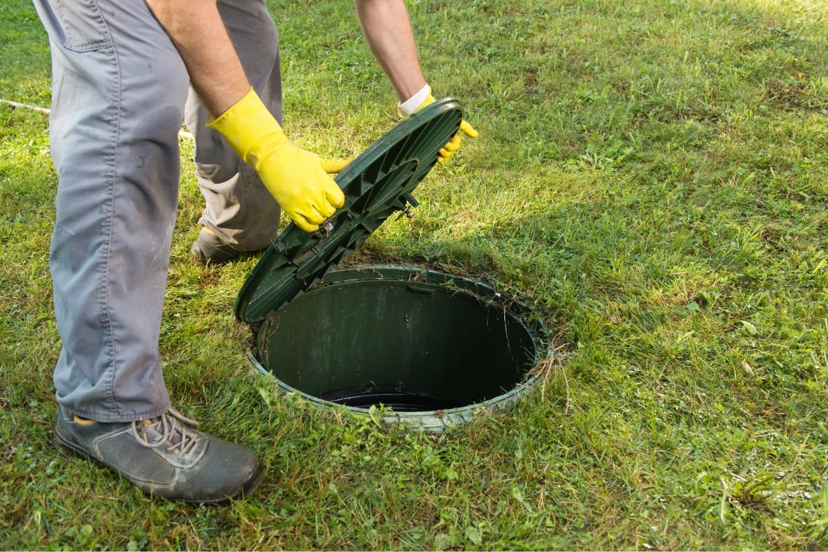 Person wearing yellow gloves lifts a green lid off a round underground septic tank on grassy ground, pondering the benefits of a mound septic system vs conventional options.