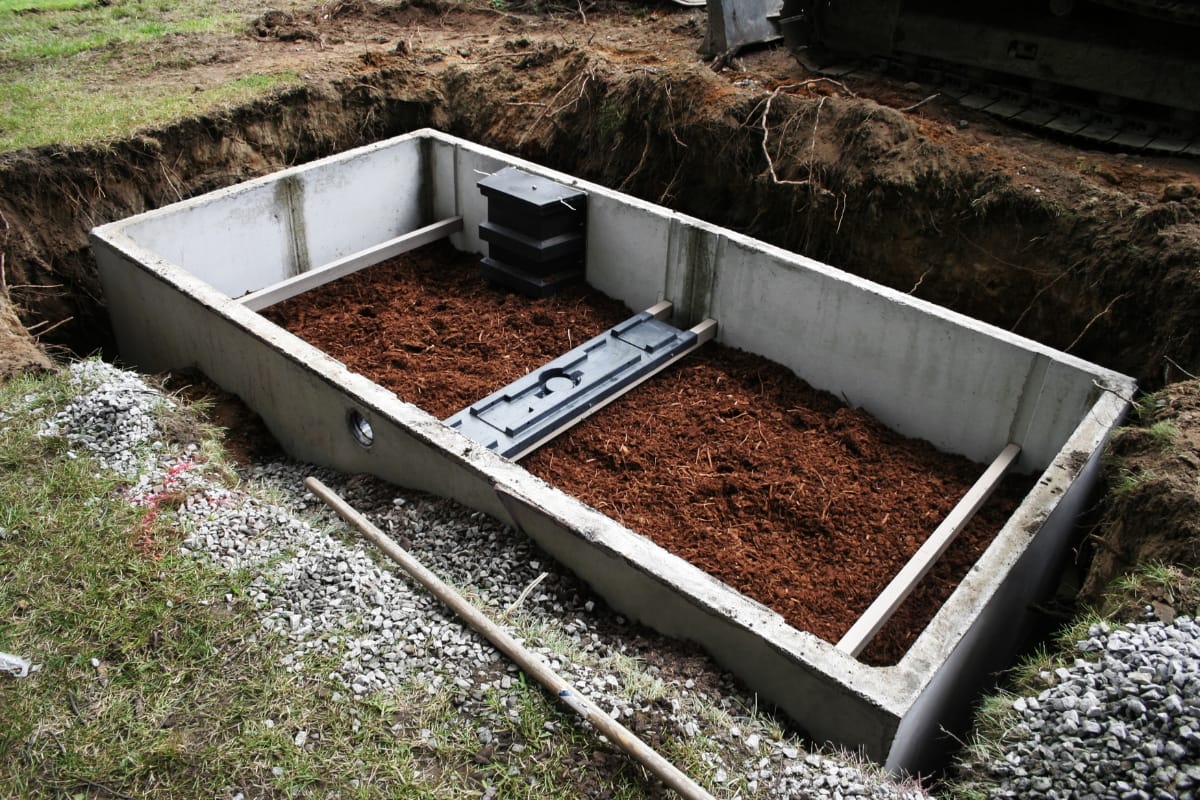 A concrete storm shelter, resembling the structure of a conventional septic system, is partially buried in the ground, with an open top revealing brown fill material inside.