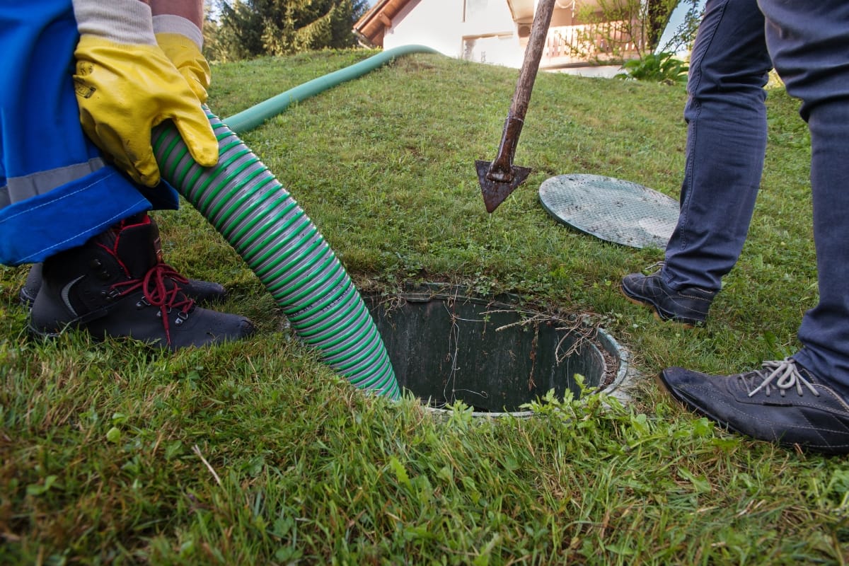 Two workers in protective gear carry out residential septic tank maintenance, efficiently draining the tank with a green hose. Nearby, a shovel and the removed tank lid rest on the grass, ready for action.