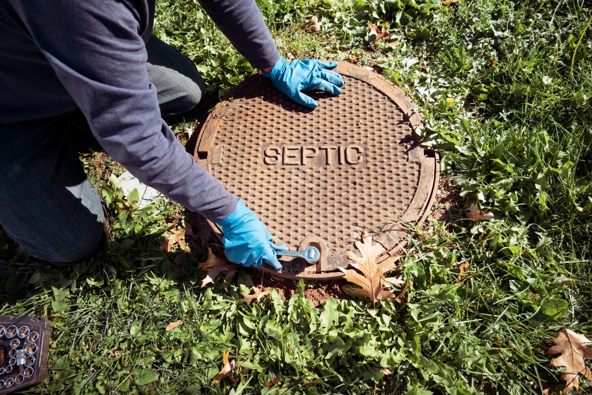 A person in blue gloves kneels on the grass, expertly using a tool to open a round septic tank cover, illustrating the careful attention needed for residential septic tank maintenance.