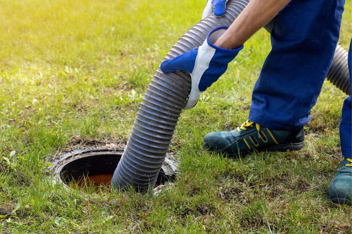 A person in gloves and protective clothing manages a hose, carefully inserting it into a ground opening on a grass surface, as part of routine residential septic tank maintenance.