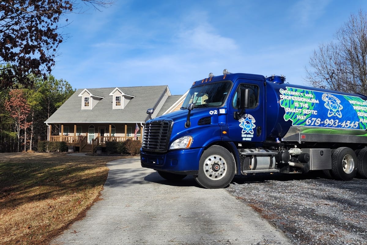 A blue septic service truck is parked on the driveway of a large house with a porch and gabled roof, surrounded by trees, ready for residential septic tank maintenance.