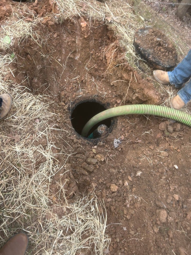 Close-up of a green hose used for residential septic cleaning inserted into an underground tank opening, surrounded by dry soil and grass, with two people standing nearby wearing work boots.