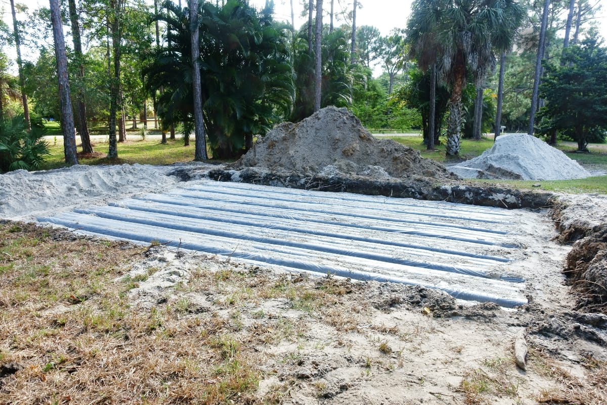 A construction site with exposed horizontal pipes in the ground hints at careful planning to meet septic drain field requirements, surrounded by piles of dirt and shaded by trees in the background.