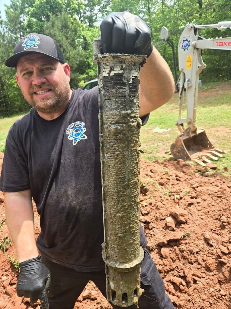 A person in a black shirt and cap holds a cylindrical object covered in mud, hinting at the labor-intensive process of residential septic cleaning. An excavator stands in the background on a dirt-covered area, ready for more work.