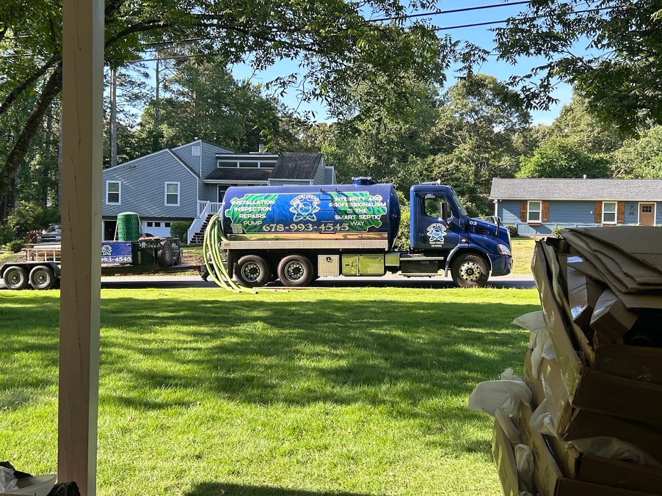 A blue septic service truck, equipped with large hoses for residential septic cleaning, is parked on a leafy street, surrounded by trees and houses.