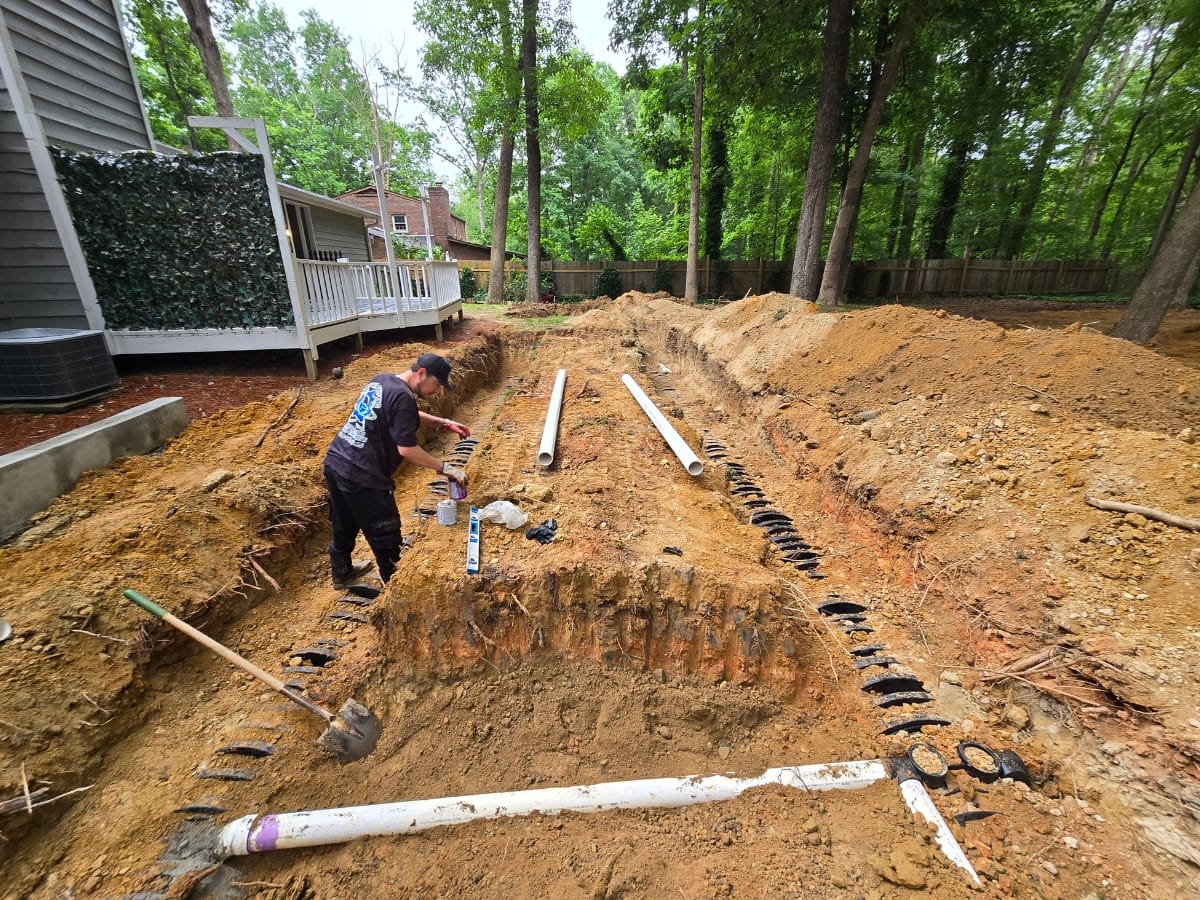 A person diligently works on a trench with exposed pipes in a backyard, implementing sustainable wastewater management practices amidst lush trees and a house in the background.