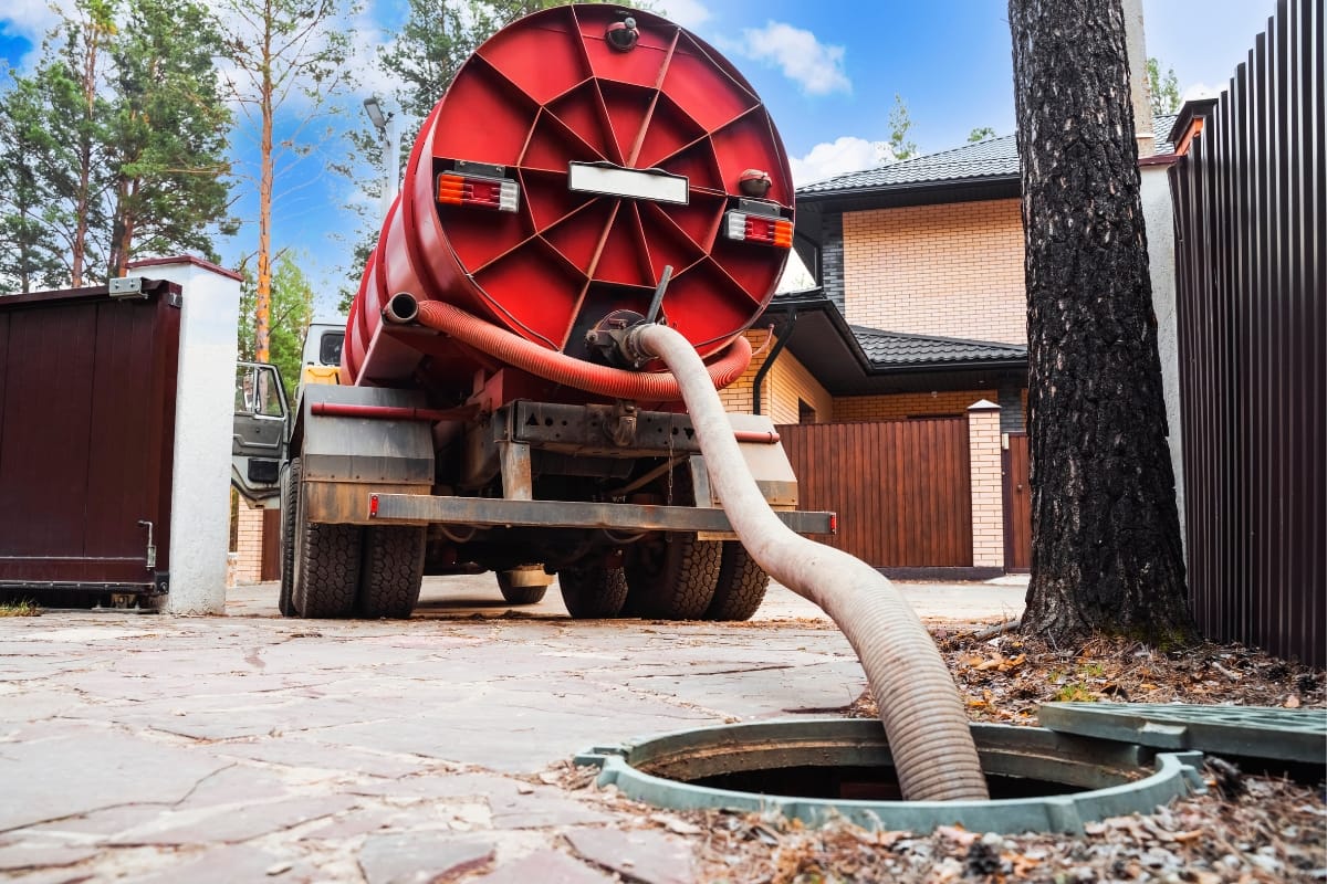 In a peaceful residential area, a septic truck with a hose inserted into an open manhole sits near a brick house, enveloped by trees. Homeowners often wonder when to pump their systems to maintain this tranquility.