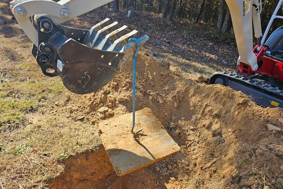 An excavator, knowing when to pump its hydraulic powers, lifts a concrete slab with a blue strap from a dirt hole in the wooded area.