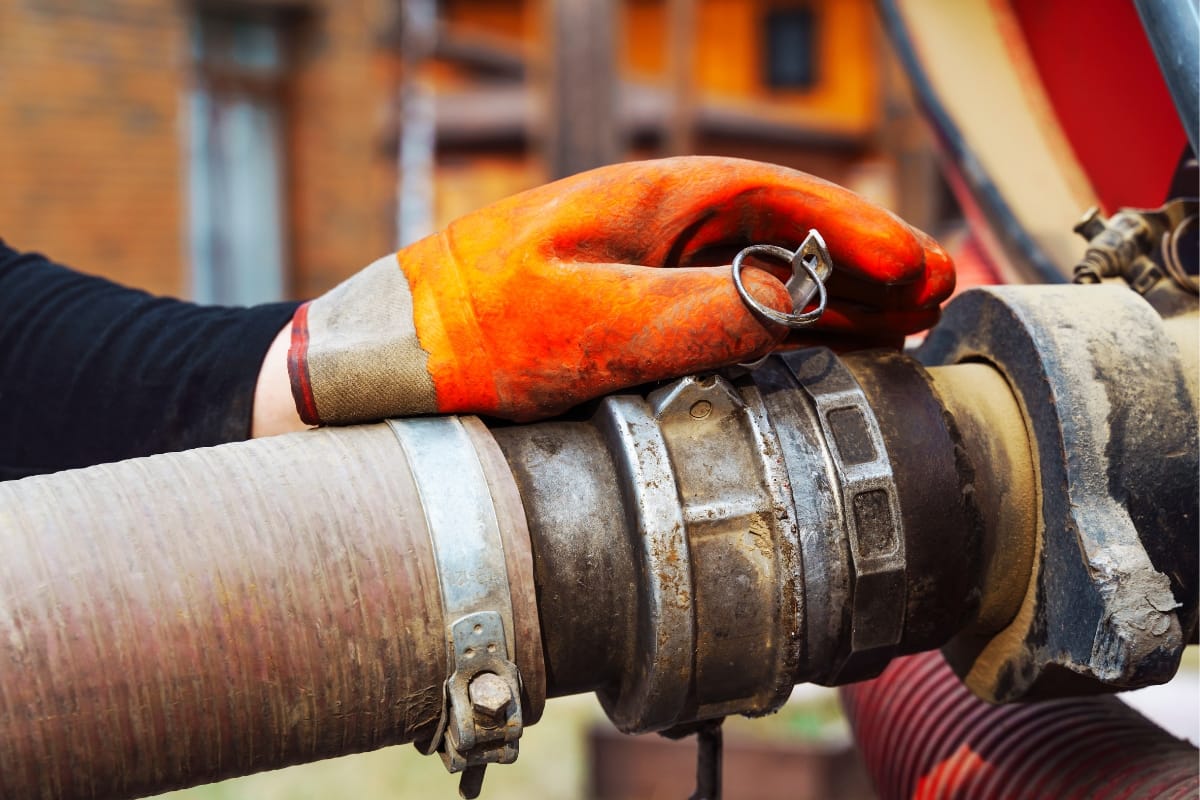 A person wearing an orange glove skillfully manages a large metal hose connector outdoors, efficiently preparing for a commercial septic tank pumping operation.