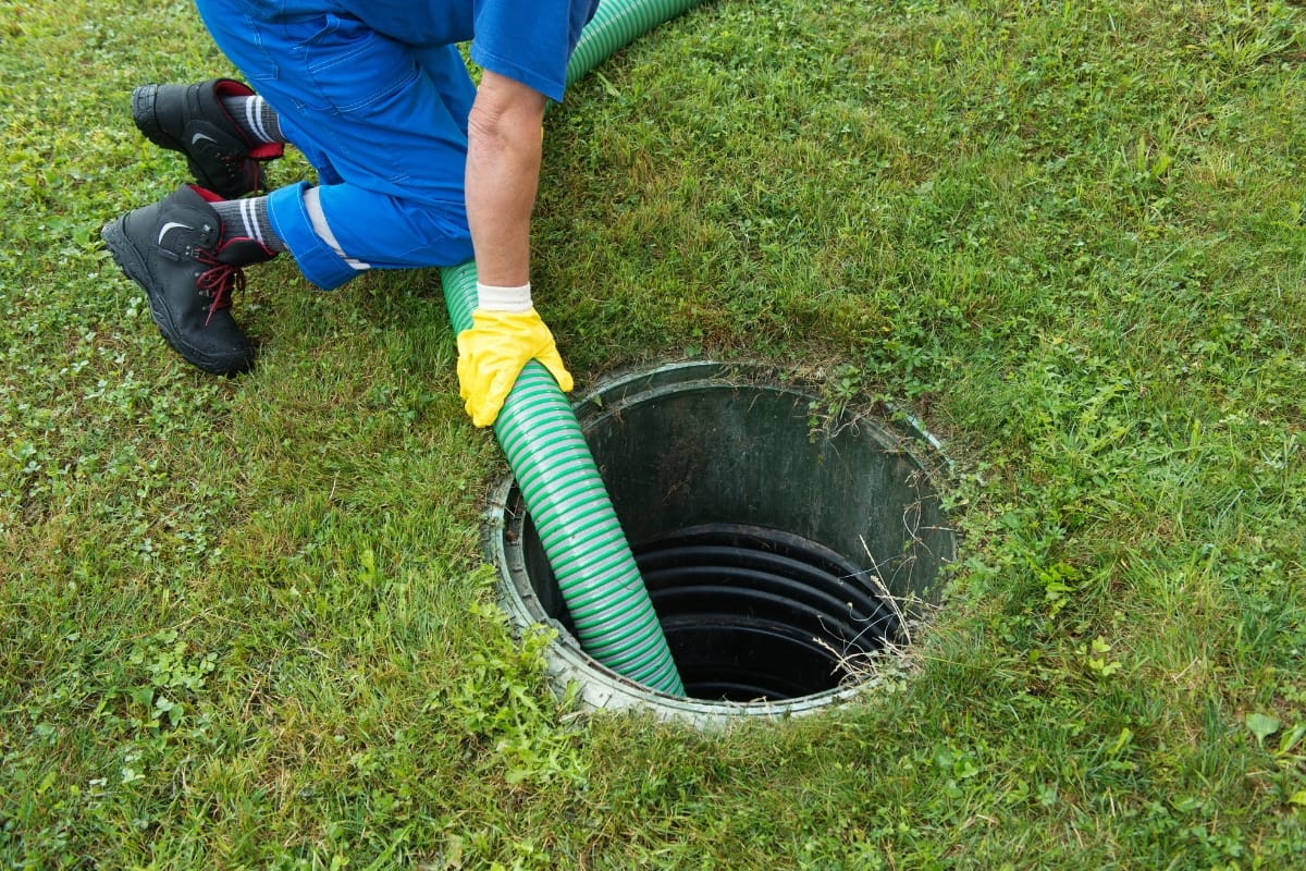 A professional in a blue uniform and yellow gloves expertly uses a hose to perform commercial septic tank pumping on a round outdoor unit set on the lush, grassy ground.