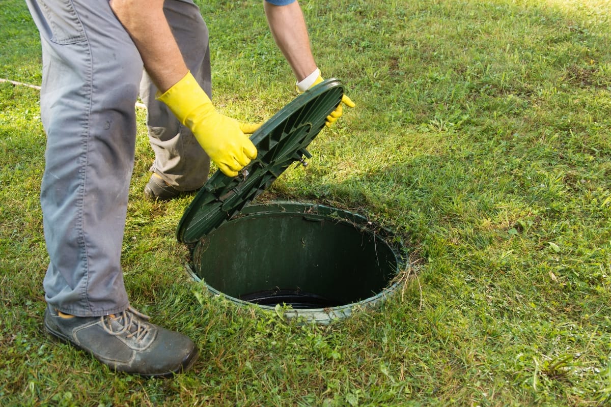 A person in gray pants and yellow gloves carefully opens a green septic tank lid on the grass, demonstrating the meticulous nature of commercial septic tank pumping.