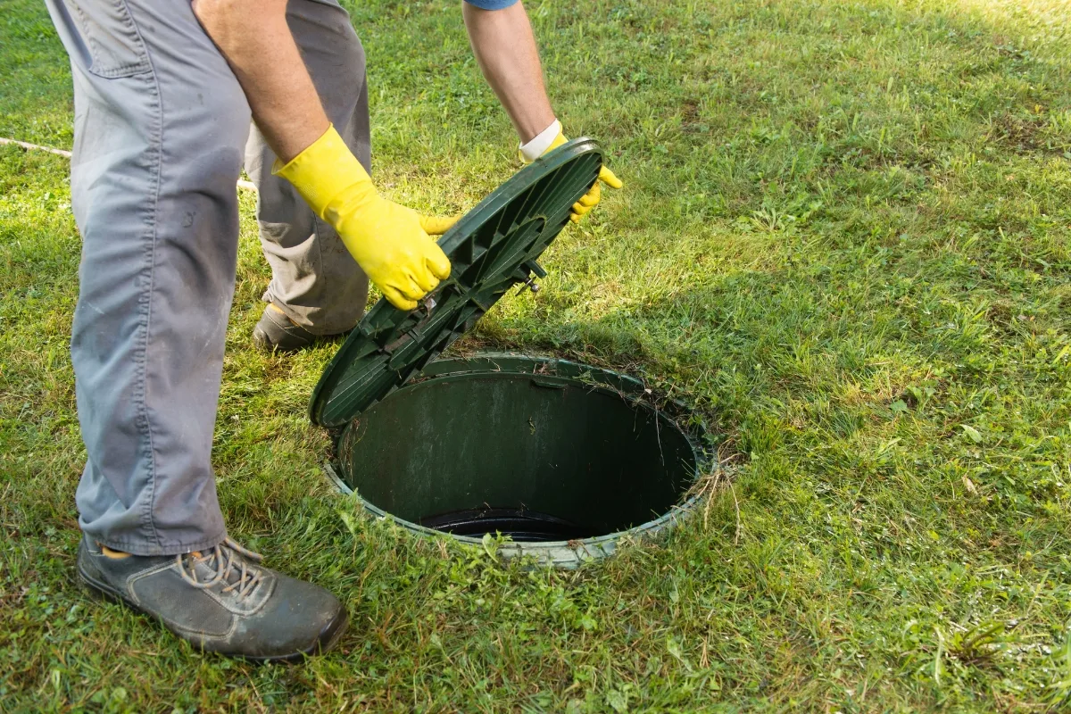 Optimal Scheduling for Commercial Septic Tank Pumping 6 A person in gray pants and yellow gloves carefully opens a green septic tank lid on the grass, demonstrating the meticulous nature of commercial septic tank pumping.
