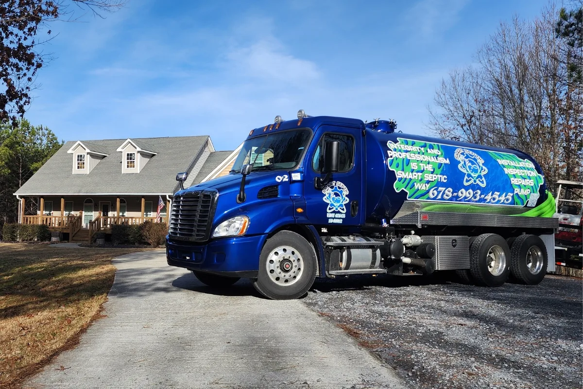 a sturdy concrete septic tank surrounded by lush vegetation, showcasing the durability and traditional appeal of this material option. A blue septic service truck, ready for emergency septic repair, parked on a residential driveway in front of a house.
