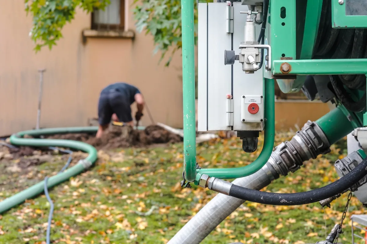 Managing Your Emergency Septic Repair Crisis: Step-by-Step Guidance 5 A person focused on emergency septic repair works diligently on the ground near a large, green industrial machine with hoses attached. Fallen leaves are scattered on the grass, adding an autumn touch to the scene.