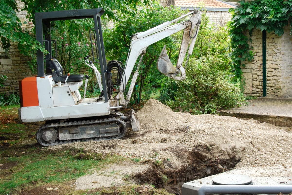 A small excavator, often used by local septic tank installers, diligently digs in a garden area next to a stone wall and trees.