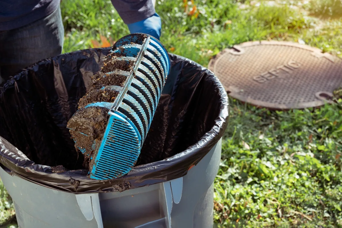 Strategies for Septic Systems to Lower Nitrogen Pollution 4 A person cleans a septic tank filter filled with sludge, holding it above a trash can outdoors, mindful of the nitrogen pollution that unchecked buildup can contribute to the environment.