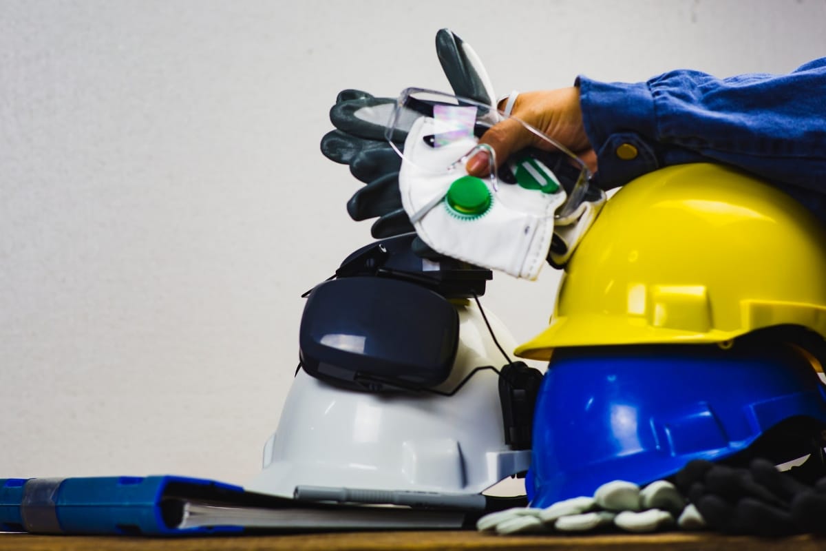 A person holds safety gear, including goggles, gloves, and masks, over a stack of white, blue, and yellow hard hats on a table with a folded safety vest nearby—perfect for anyone about to replace a septic tank lid.