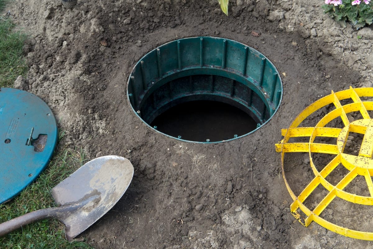 A ground hole surrounded by a green rim, featuring a yellow circular metal grate, hints at the task ahead—replacing the septic tank lid—with a shovel conveniently nearby for the job.