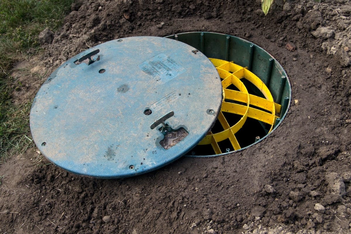 A partially open blue manhole cover, designed to replace a septic tank lid, reveals yellow interior components set into the ground, surrounded by soil.