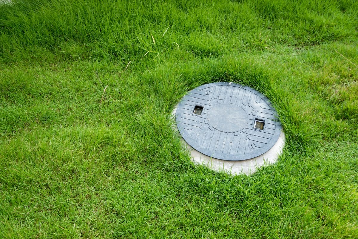 A round manhole cover, resembling the style needed to replace a septic tank lid, is surrounded by lush green grass.